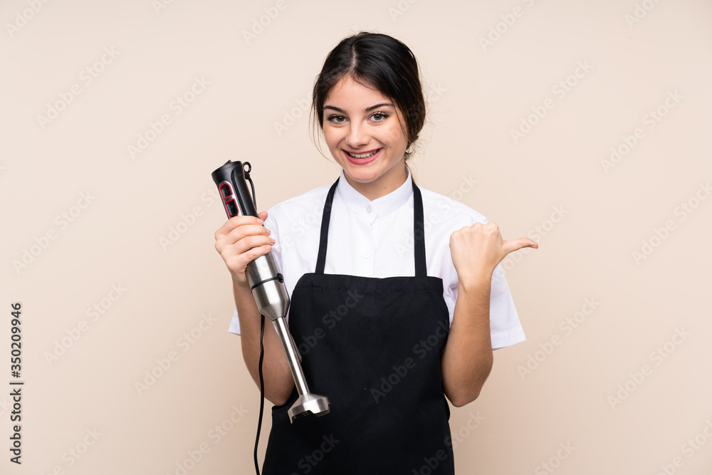 Young woman using hand blender over isolated background pointing to the side to present a product