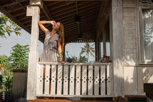 Young beautiful woman relaxing on cozy wooden balcony. Attractive girl sit on open-air veranda of wooden bungalow with tropical garden view. Summer vacation on Bali island. Countryside lifestyle.