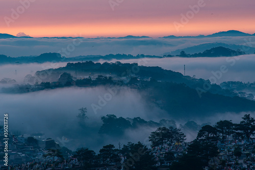 Mountains in fog at beautiful morning in autumn in Dalat city  Vietnam. Landscape with Langbiang mountain valley  low clouds  forest  colorful sky   city illumination at dusk.