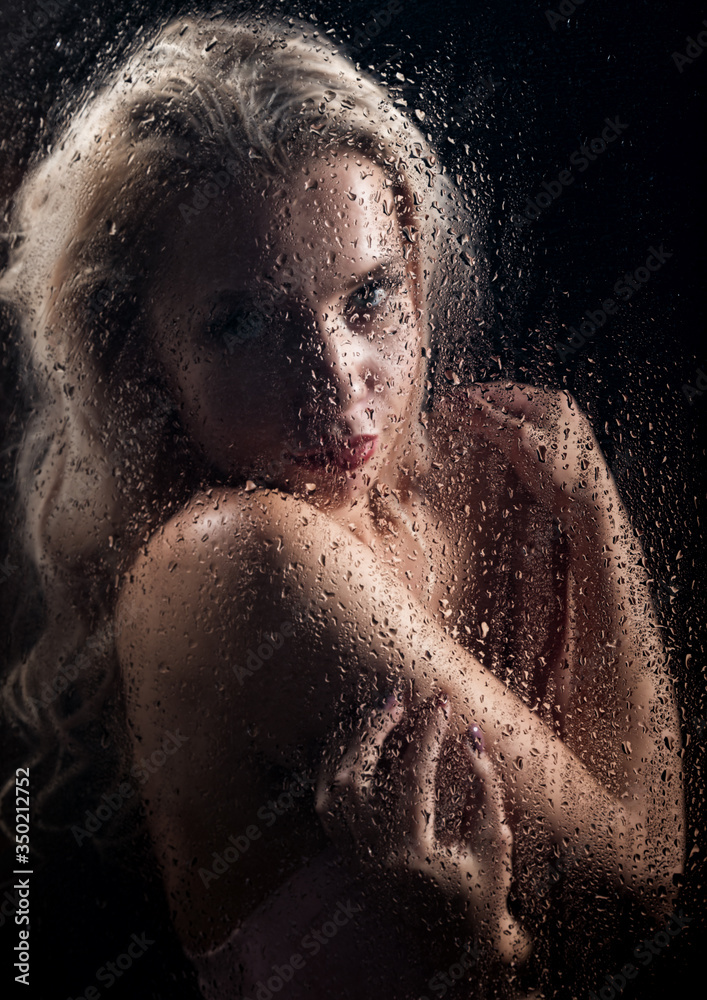beautyful blonde woman behind wet glass on a dark background, woman takes a shower