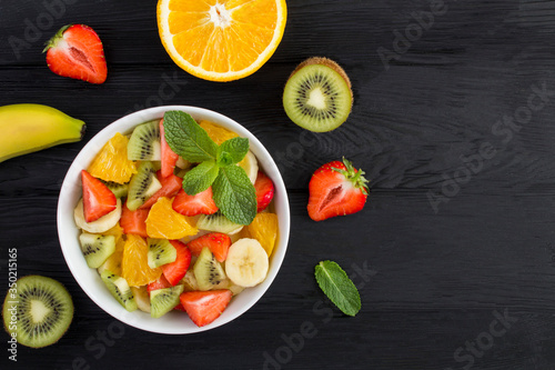 Fruit salad in the white bowl and ingredients on the black wooden background. Top view. Copy space.
