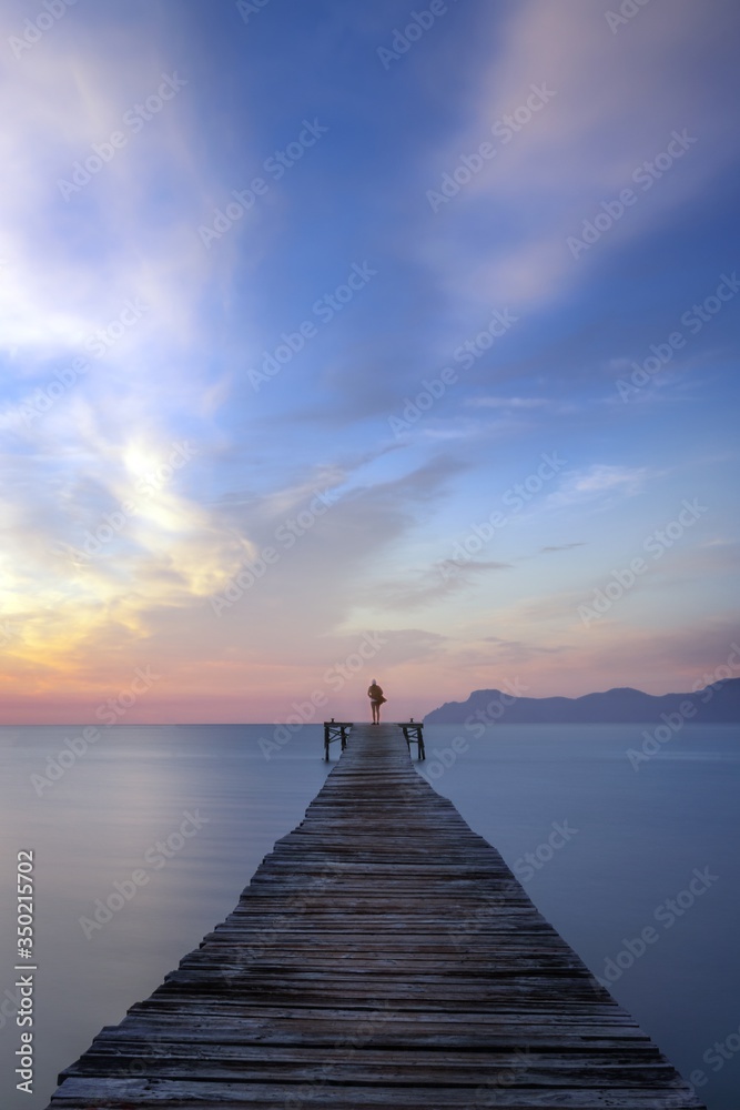 Fototapeta premium Lone person standing at end of wooden boardwalk at sunrise, with calm sea and beautiful colourful sky and mountains in the distance, Alcudia beach, Mallorca, Spain.