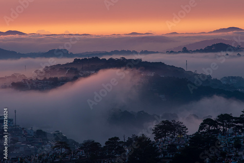 Mountains in fog at beautiful morning in autumn in Dalat city, Vietnam. Landscape with Langbiang mountain valley, low clouds, forest, colorful sky , city illumination at dusk. © Nhan