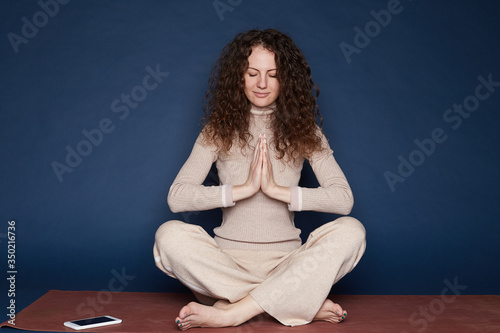 Optimistic female meditates indoors, tries keep mind clear, has minute relaxation after work, keeps calm inside of soul, knows technique for rest, wears casual beige outfit, isolated on blue wall.