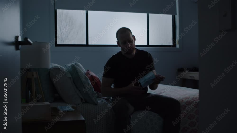 Man Sitting on Bed in Dark Room Puts Face Mask on. Wide shot Pushes In Man Sitting on Bed in Dark Room Puts Face Mask on. Wide shot Pushes In