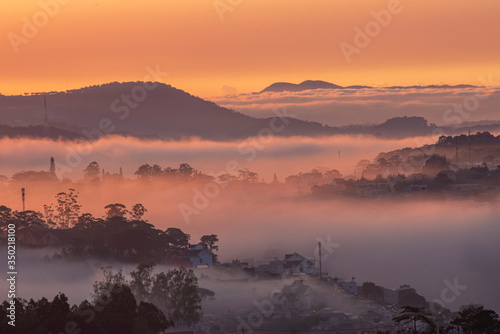 Mountains in fog at beautiful morning in autumn in Dalat city, Vietnam. Landscape with Langbiang mountain valley, low clouds, forest, colorful sky , city illumination at dusk. © Nhan