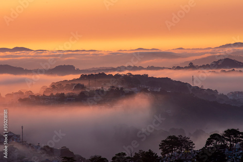 Mountains in fog at beautiful morning in autumn in Dalat city, Vietnam. Landscape with Langbiang mountain valley, low clouds, forest, colorful sky , city illumination at dusk.