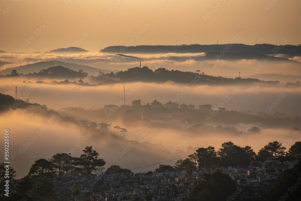 Mountains in fog at beautiful morning in autumn in Dalat city, Vietnam. Landscape with Langbiang mountain valley, low clouds, forest, colorful sky , city illumination at dusk.