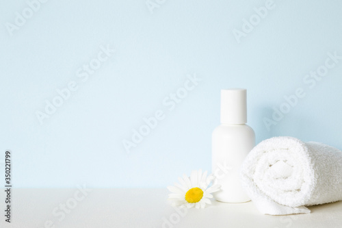 White bottle and towel on shelf. Beautiful chamomile or daisy. Fresh flower. Care about clean and soft face, hands, legs and body skin. Empty place for text on pastel blue wall. Front view.