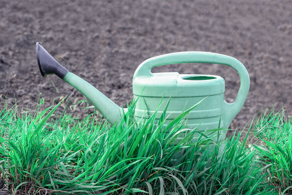 Green plastic watering can close up standing on grass with farm/garden land behind. Farming, gardening concept Green plastic watering can close up standing on grass with farm/garden land behind. Farming, gardening concept