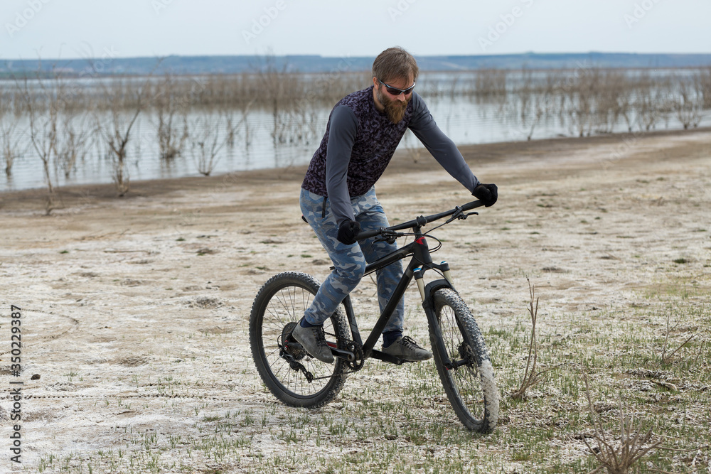 Fototapeta premium Sports brutal bearded guy on a modern mountain bike. A cyclist in a salt deserted place by the lake.