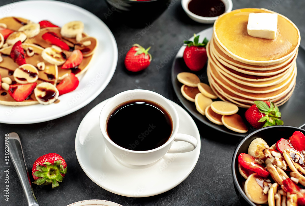 The concept of a delicious breakfast. Coffee and mini pancakes with strawberries, bananas, nuts in a plate and American pancakes in a plate on a stone background The concept of a delicious breakfast. Coffee and mini pancakes with strawberries, bananas, nuts in a plate and American pancakes in a plate on a stone background