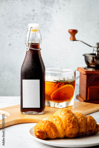 Iced cold brew coffee in blank label bottle on marble table decoration with ice ball in whisky glass, crossaint, grinder and rustic white background. vertical image and main composition on center