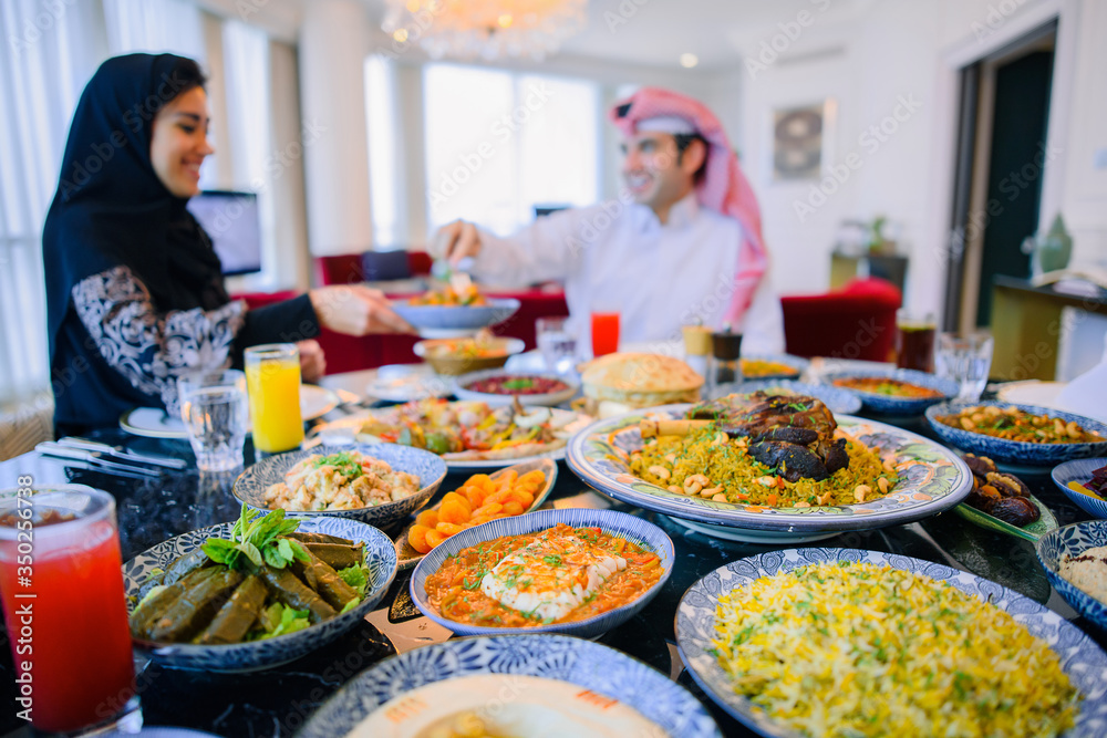 Arab family sitting on a background of a full table of food. A man in white traditional clothes and a woman in black abaya chatting at home before Iftar Arab family sitting on a background of a full table of food. A man in white traditional clothes and a woman in black abaya chatting at home before Iftar