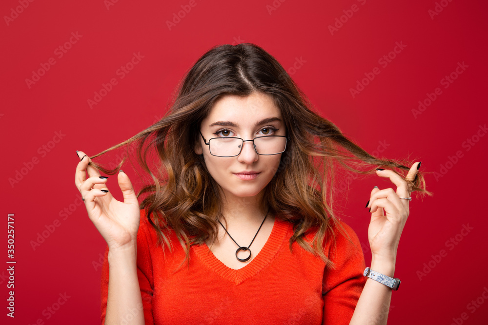 The girl in glasses twists hair with fingers on a red background. The girl in glasses twists hair with fingers on a red background.