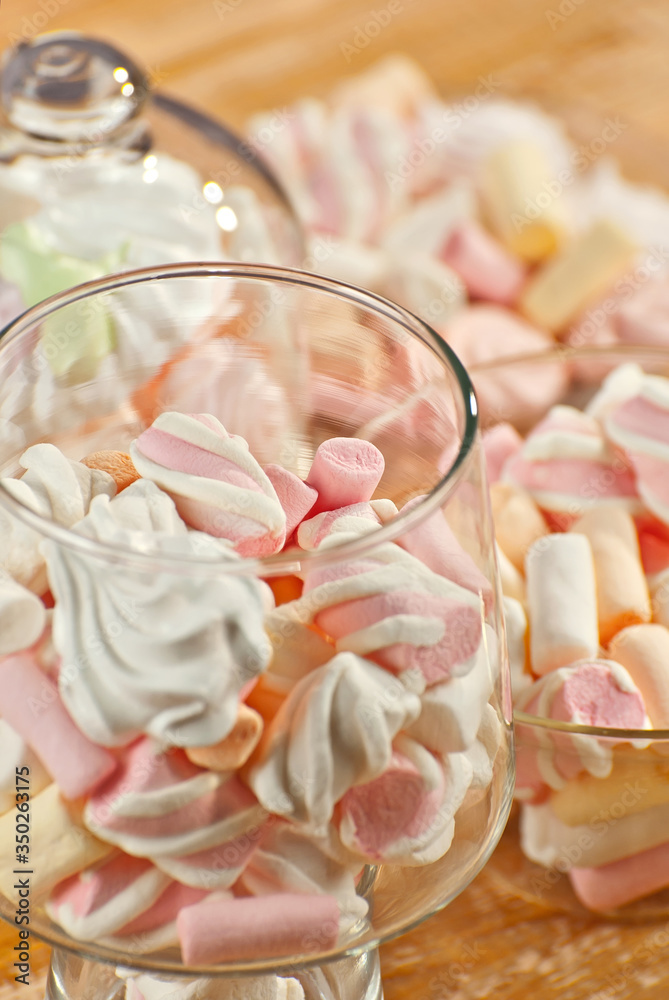 Sweets in a glass on a wooden table. Transparent utensils with marshmallows and biz on a shabby old board. Pink with white soft candies close up.