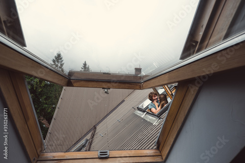 A man and a woman look out of the windows of a house in the mountains.