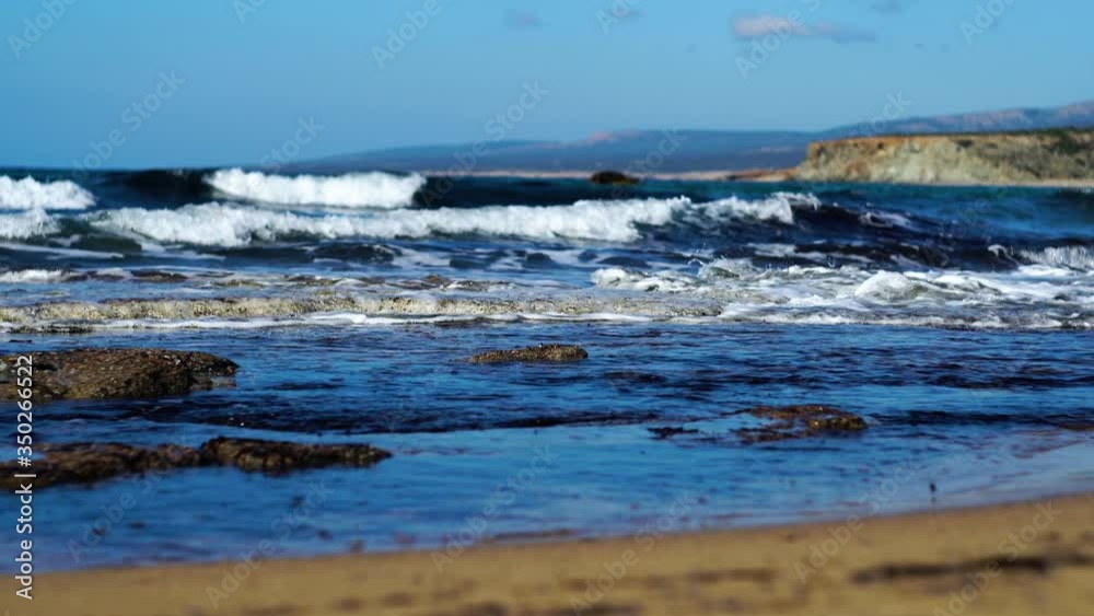 Dark blue sea waves splashing on sandy beach on sunny summer day. Foamy waters of Mediterranean Sea crashing on rocks and rolling on sand. Tropical paradise, beauty in nature, seascape.