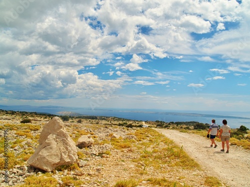 Typical czech tourists walking on tour in dry stony landscape on the top of hill on island Rab with sea view and clouds over horizon during summer holidays, active vacation