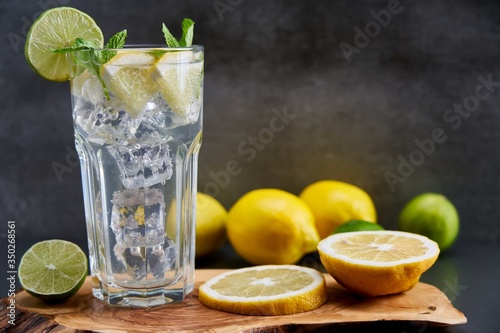 Cold refreshing limonade  mojito or gin tonic in glass  with fresh mint and ice cubes  lime and lemon on wooden board and black background. Summer cocktails and drinks concepts. Copy space for text.