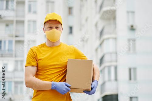 Delivery Man employee in yellow uniform cap, t-shirt, face mask and gloves holds a cardboard box package on building backdrop. Safety delivery quarantine service in covid-19 virus pandemic period.