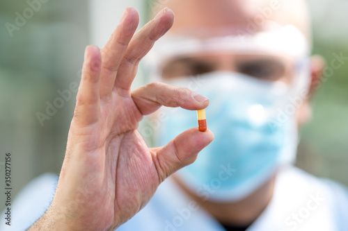 Out of focused doctor wearing surgical mask and medical safety goggles holding a medication pill in his fingers