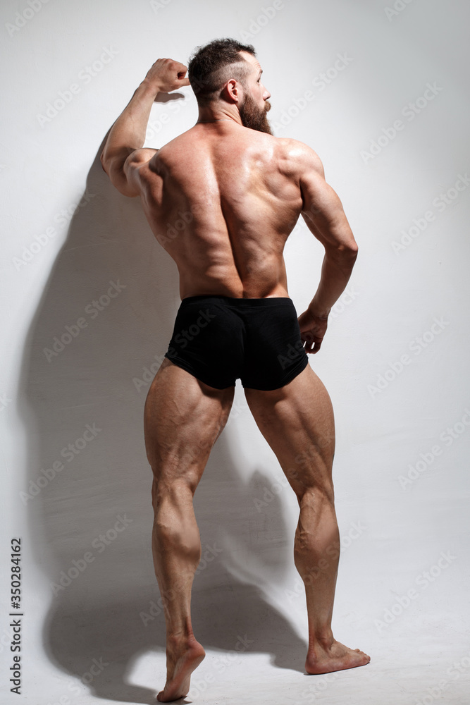 Athletic bearded man shows muscles standing in full growth on a light background view from the back.