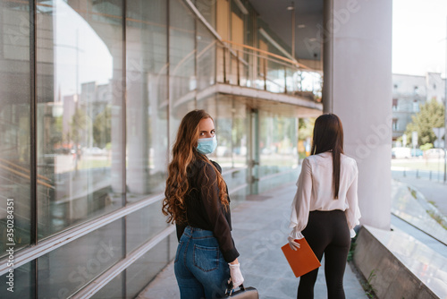 Two beautiful attractive caucasian business women with a mask and gloves stand in front of the company. COVID - 19 virus protection