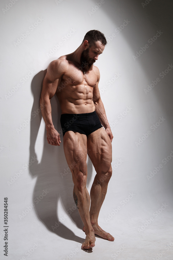 Athletic bearded man shows muscles standing in full growth on a light background.