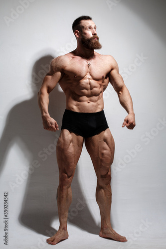 Athletic bearded man shows muscles standing in full growth on a light background.