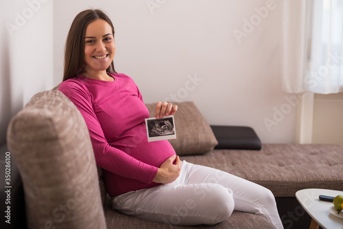 Pregnant woman sitting on the sofa and holding ultrasound picture. 