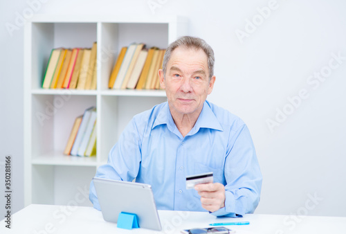 Senior citizen man working on a tablrt holding glasses on his forehead looking at a credit card in his hand photo