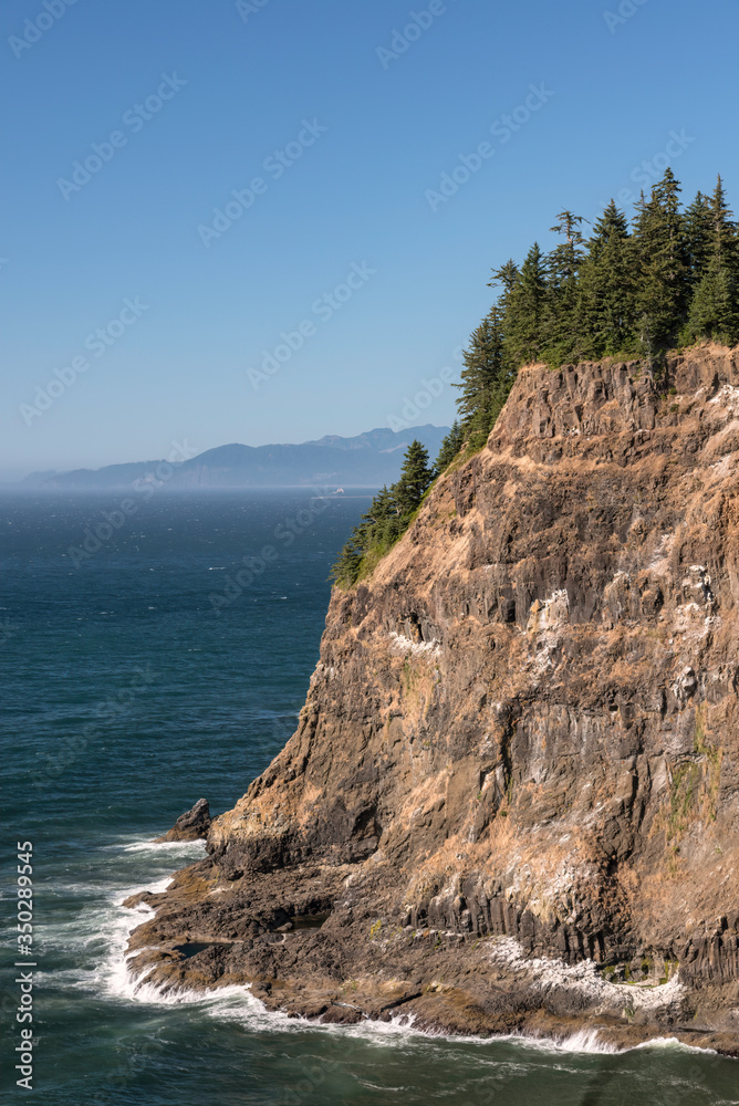 Steep cliff on the Oregon coast surrounded by waves with fog in the background.