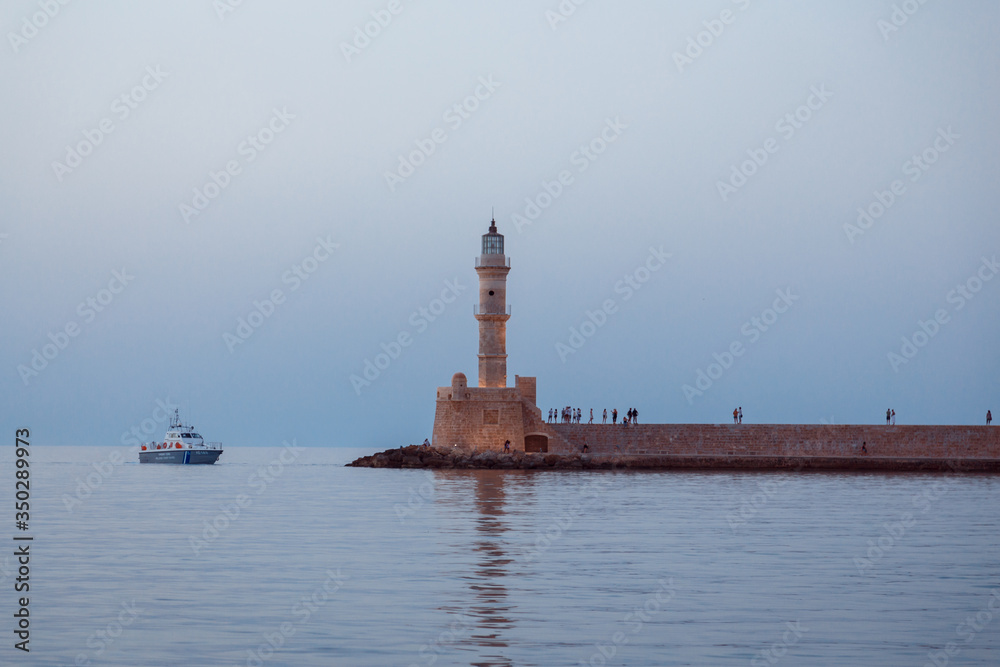 lighthouse on the water. People walk on the pavement. A small passenger boat passes nearby. Romantic atmosphere of a European evening.