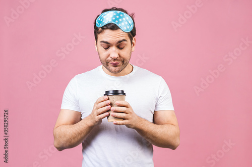 Portrait of nice calm peaceful sleepy young man with blue sleeping mask and cup of coffee or tea. Isolated over pink background. © denis_vermenko