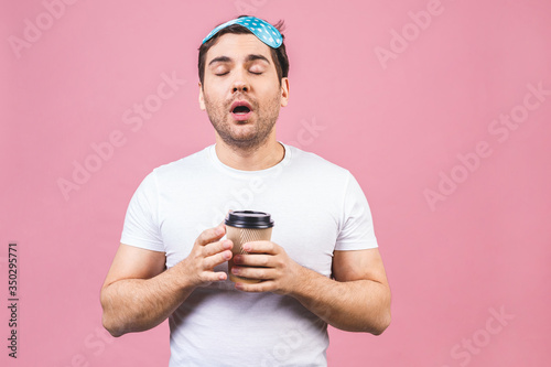 Portrait of nice calm peaceful sleepy young man with blue sleeping mask and cup of coffee or tea. Isolated over pink background. © denis_vermenko