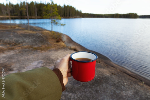 Male hand holding a mug of a hot tea on the background of the lake. Red cup with a drink in a hand.