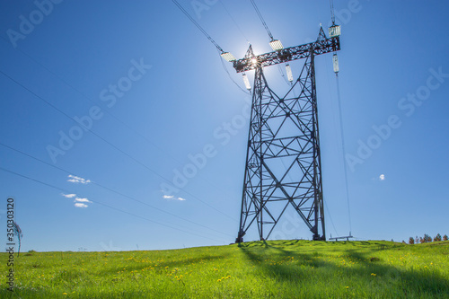metal support of a high voltage power line on top of a green hill in the backlight of the sun