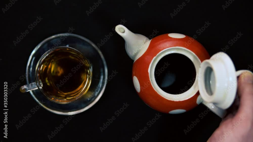 Tea ceremony. A male hand opens the ceramic lid of a hot red polished white ceramic teapot next to a glass bowl for a bowl. Black background, oncoming light 4k Tea ceremony. A male hand opens the ceramic lid of a hot red polished white ceramic teapot next to a glass bowl for a bowl. Black background, oncoming light 4k