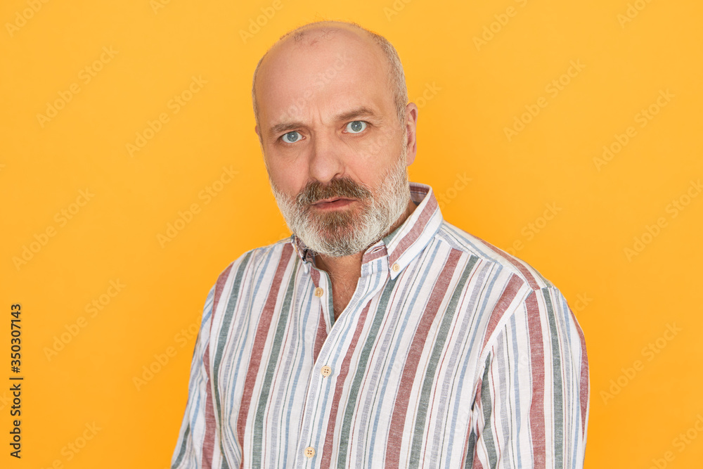 Obraz premium Horizontal shot of grumpy displeased senior man with thick gray beard and bald head staring at camera having suspicious look, frowning, not trusting you. Human facial expressions and reaction