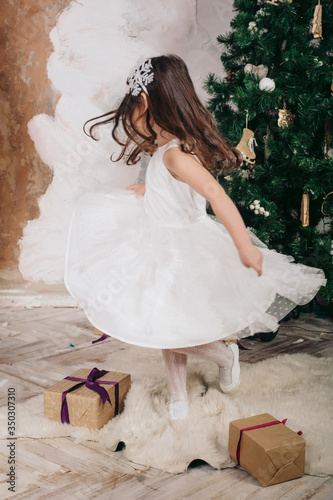 a little girl in a dress dances near the Christmas tree, on the floor there are gifts in boxes photo