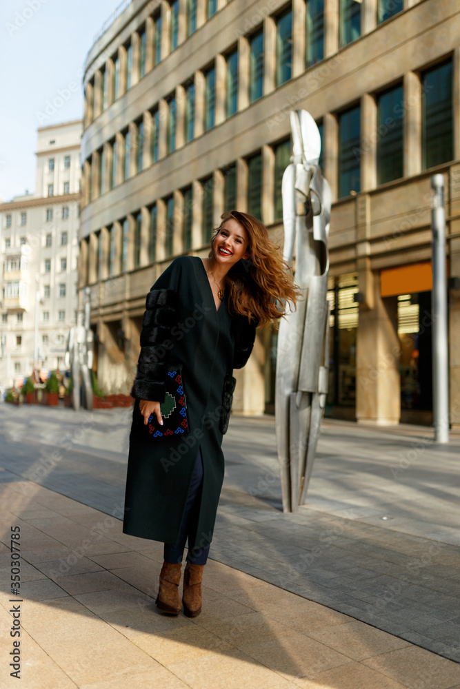 Fashion outdoor photo of happy young woman with red hair in elegant clothes and luxurious coat, walking by the city.