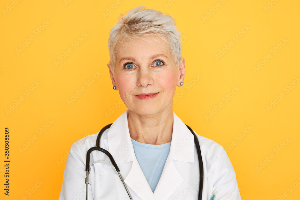 Isolated image of successful middle aged woman physician with short blonde dyed hair having medical consultation, studying current concerns and symptoms, posing against yellow wall background Isolated image of successful middle aged woman physician with short blonde dyed hair having medical consultation, studying current concerns and symptoms, posing against yellow wall background