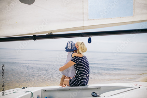 Mom and her little daughter sit on a yacht, little girl hugs her mother, they look into the distance at the sea