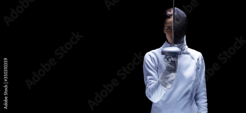 Charming girl in a fencer costume posing on a black background. The sword separates the head into a face and a mask. The concept of fencing. photo