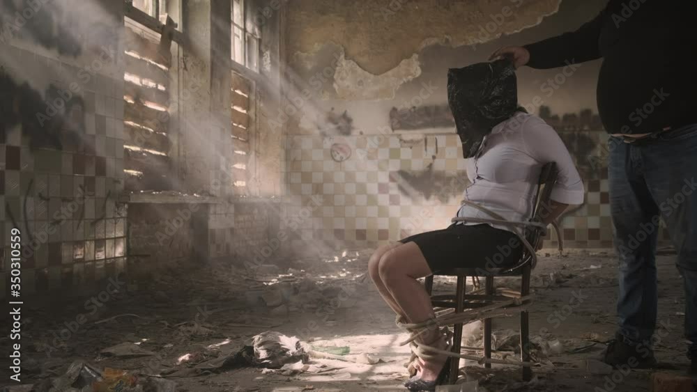 female hostage in captivity in criminal tied with rope to chair. Kidnapper tears packet from head of kidnapped victim.