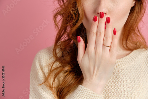 Close frame of a woman's hand with a red varnish covering her mouth. Red-haired girl on a pink background