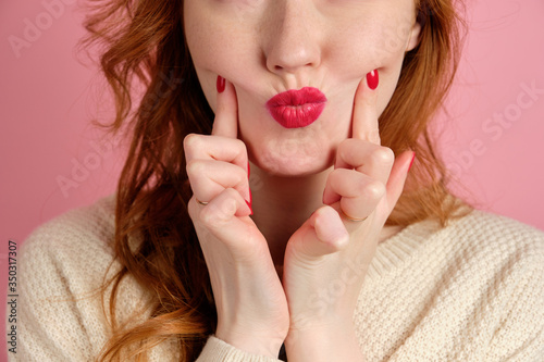 The red-haired girl pulls red lips forward and presses her fingers on her cheekbones. Close frame of lips.