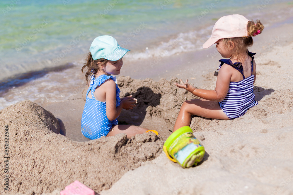 Two sister girls in swimsuits and caps playing on the beach with toys and sand on a sunny summer day. Fun family vacation at the sea. Activity outdoors Two sister girls in swimsuits and caps playing on the beach with toys and sand on a sunny summer day. Fun family vacation at the sea. Activity outdoors