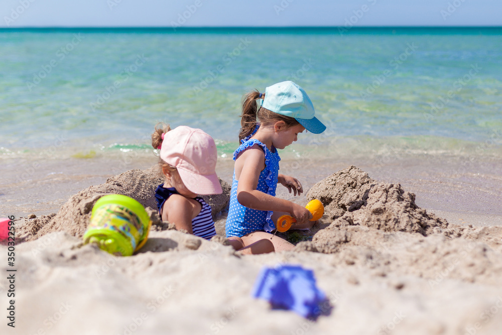 Two sister girls in swimsuits and caps playing on the beach with toys and sand on a sunny summer day. Fun family vacation at the sea. Activity outdoors Two sister girls in swimsuits and caps playing on the beach with toys and sand on a sunny summer day. Fun family vacation at the sea. Activity outdoors